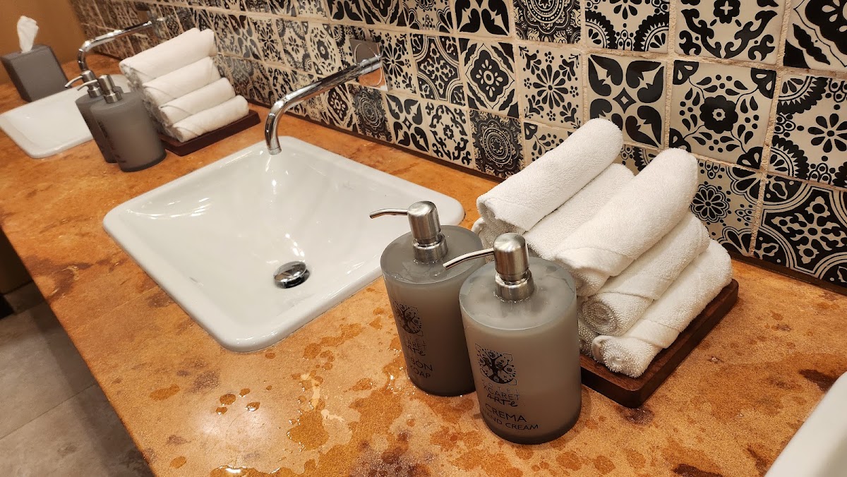 Hotel Xcaret Arte bathroom vanity with white sink, rolled towels, soap dispensers, and black patterned tile backsplash on terracotta countertop.