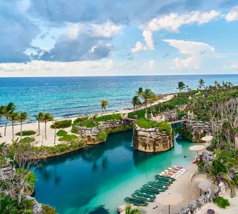 Aerial view of Hotel Xcaret Arte's beachfront lagoon with turquoise water, limestone cliffs, palm trees, and moored boats along the sandy shore.