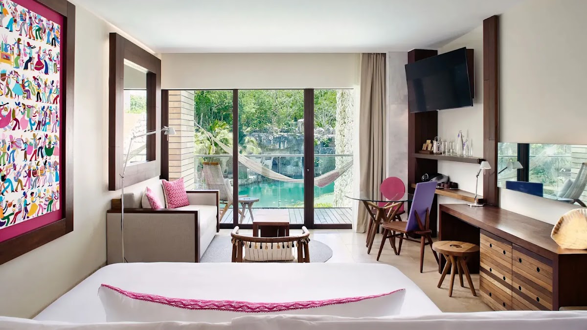 Modern resort suite with bed in foreground, living area, and glass doors opening to pool and jungle views at Hotel Xcaret Arte.