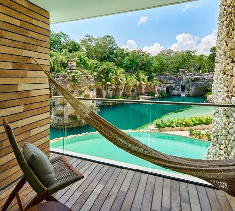 A wooden hammock sways over a cenote surrounded by lush jungle at Hotel Xcaret Arte, with Mayan ruins visible in the distance.