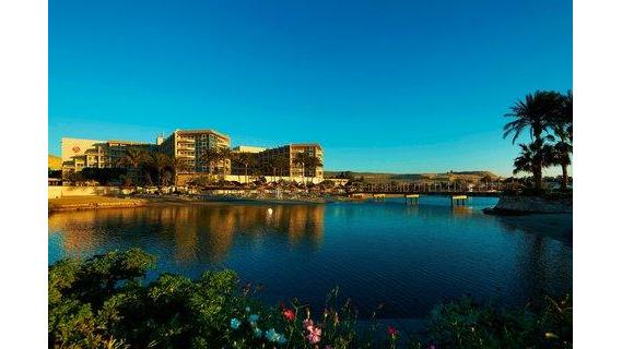 Hurghada Marriott Beach Resort's multi-story buildings overlook a calm lagoon with palm trees and flowering plants in the foreground under clear blue sky.
