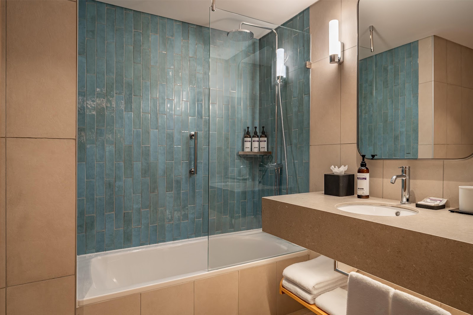 Modern bathroom with curved sink, illuminated mirror, green tile shower enclosure, and stacked towels on wooden shelf.