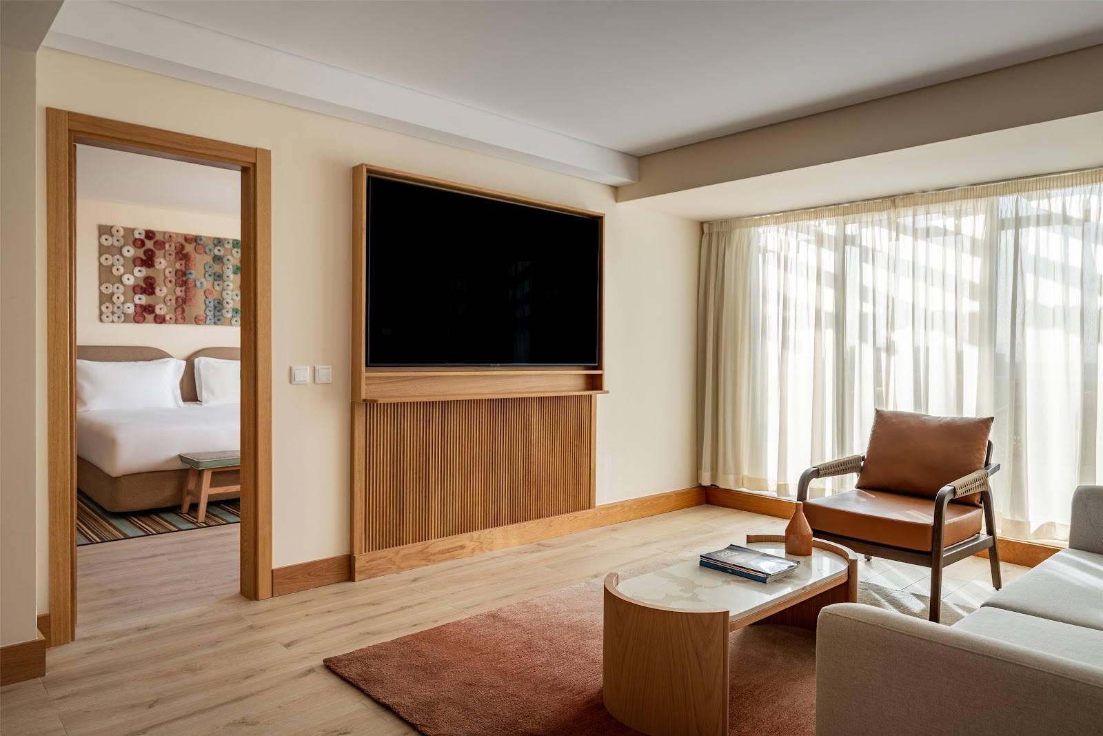 Resort suite living area with wood furnishings, flat-screen TV, leather armchair, and open bedroom doorway showing white bedding.