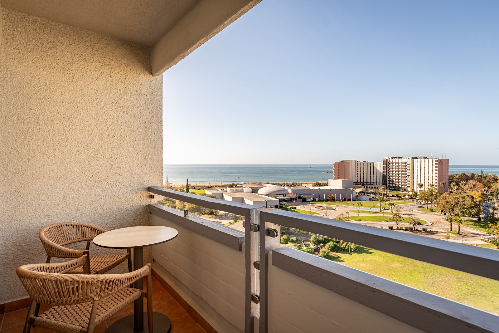 Two wicker chairs and a small table on a balcony overlooking the Algarve coastline and resort buildings.