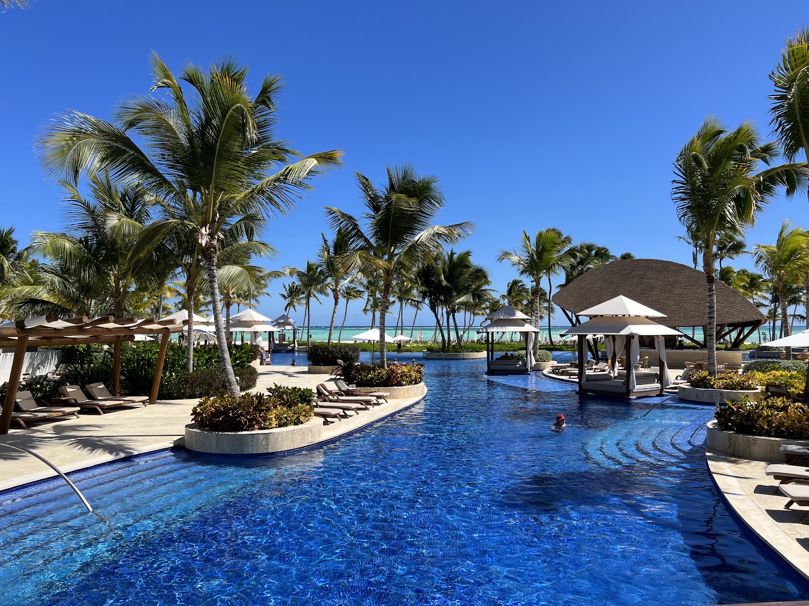 Resort pool with swim-up bar gazebo, lounge chairs under umbrellas, palm trees, and ocean view beyond under clear blue sky.