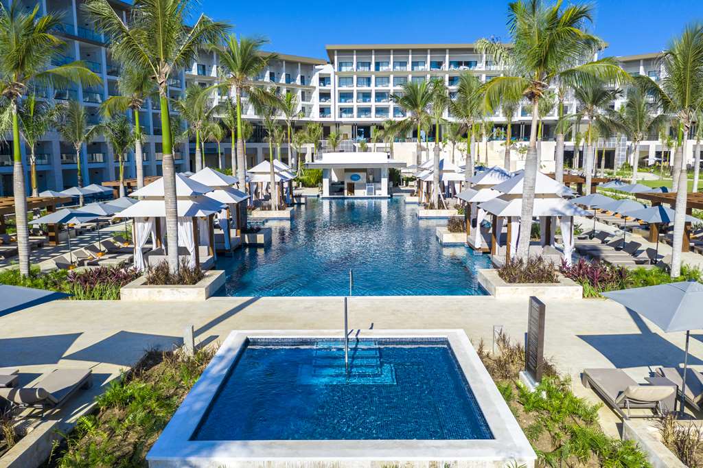 Large curved pool with a wooden-roofed palapa bar, palm trees, and white resort buildings under clear blue sky.