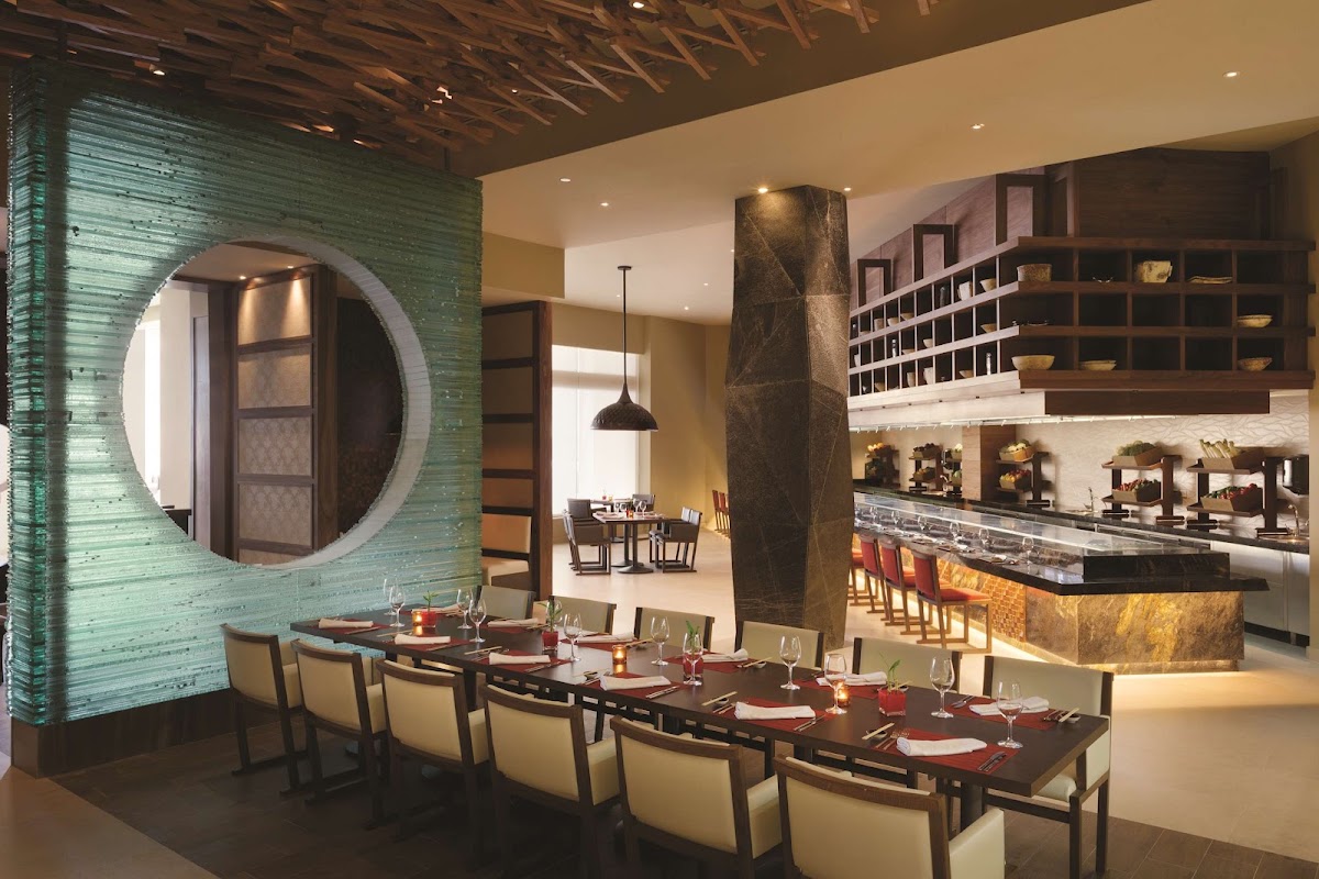 Contemporary dining room with long table set for service, green tile accent wall with round mirror, bar counter, and wooden ceiling beams.