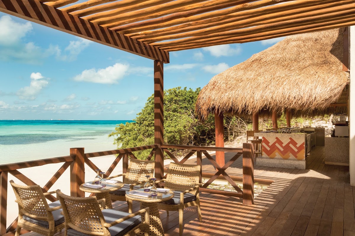 Wooden pergola shades a beachfront dining terrace with woven chairs overlooking turquoise water and white sand.