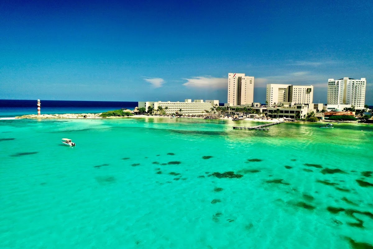 Turquoise shallow water with visible sea grass and a small boat near multi-story resort buildings and lighthouse on the sandy shore under blue sky.