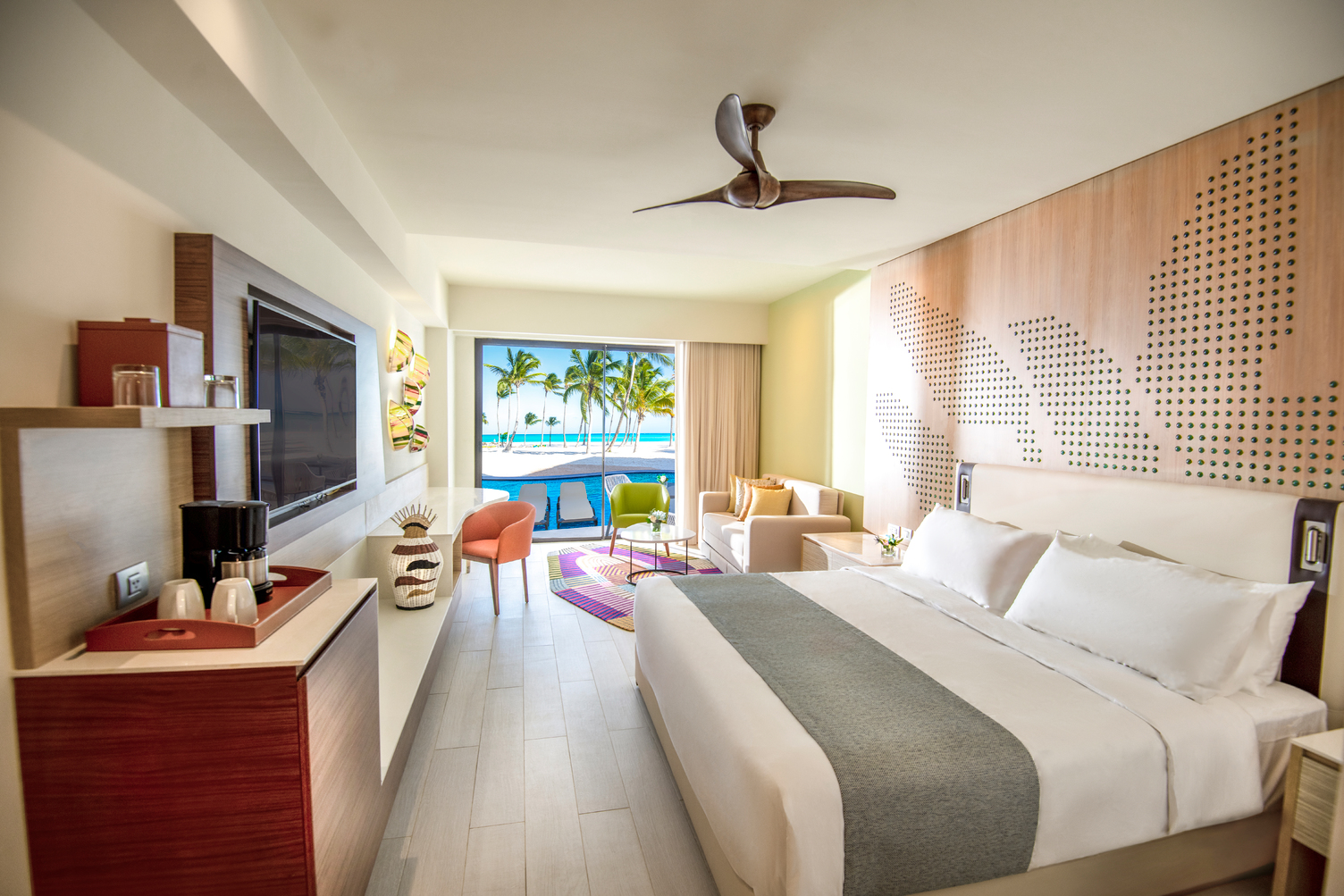 Resort guest room with king bed, wood accent wall, open balcony overlooking tropical grounds, and sitting area with orange chair.