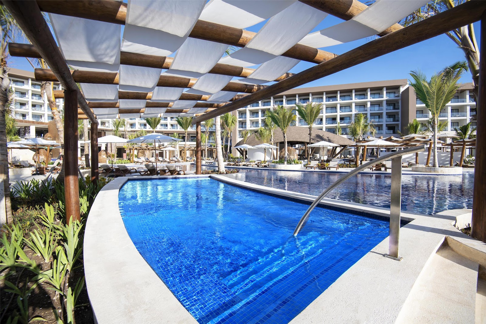 Blue swimming pool under wooden pergola with white shade sails, surrounded by palm trees and multi-story resort buildings at Hyatt Ziva Cap Cana.