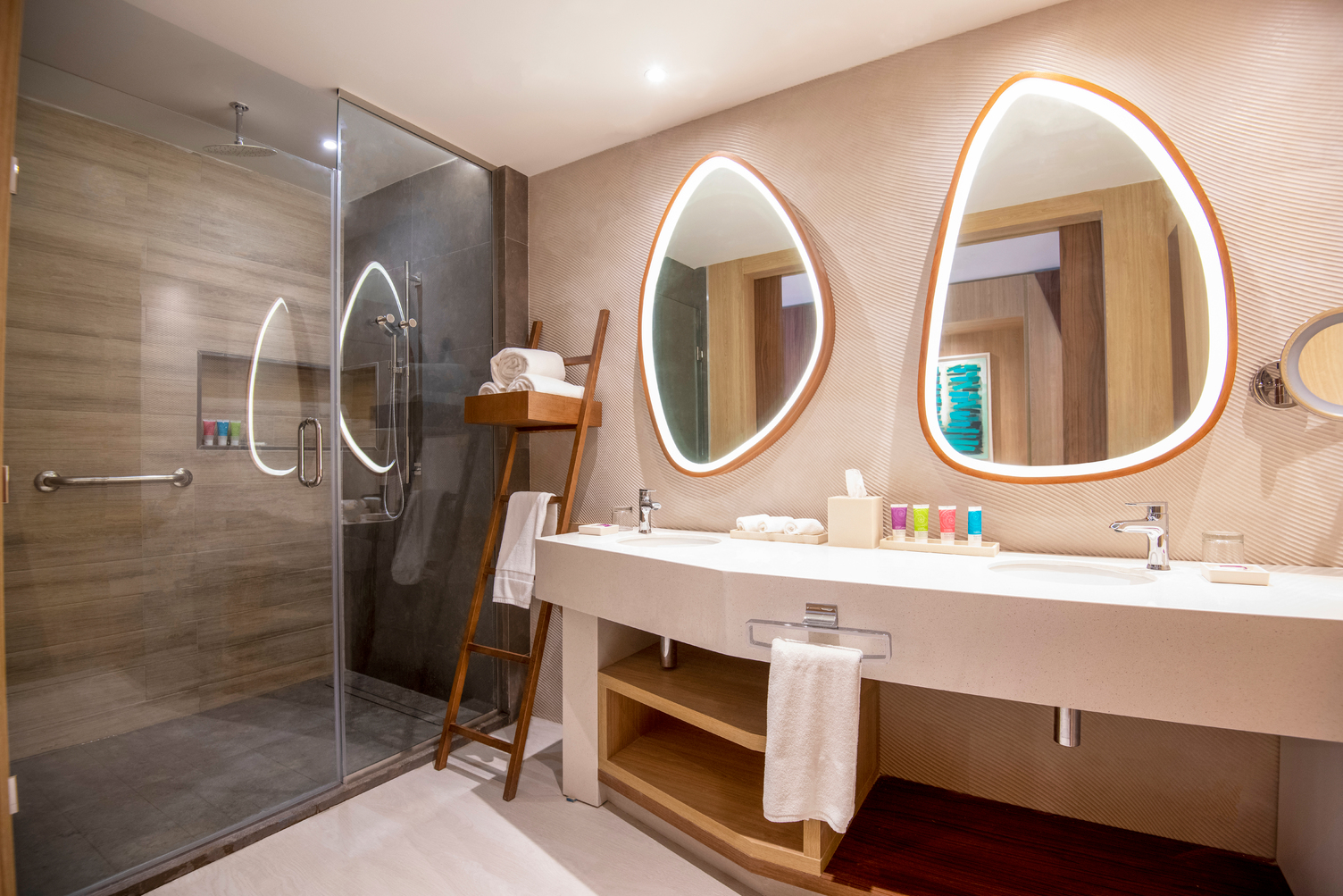 Resort bathroom with double vanity, illuminated teardrop mirrors with orange frames, walk-in shower, and wooden ladder towel rack.