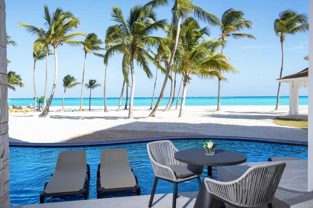 Man wading in bright blue pool toward a cabana with white curtains and palm trees under clear sky at Hyatt Ziva Cap Cana.