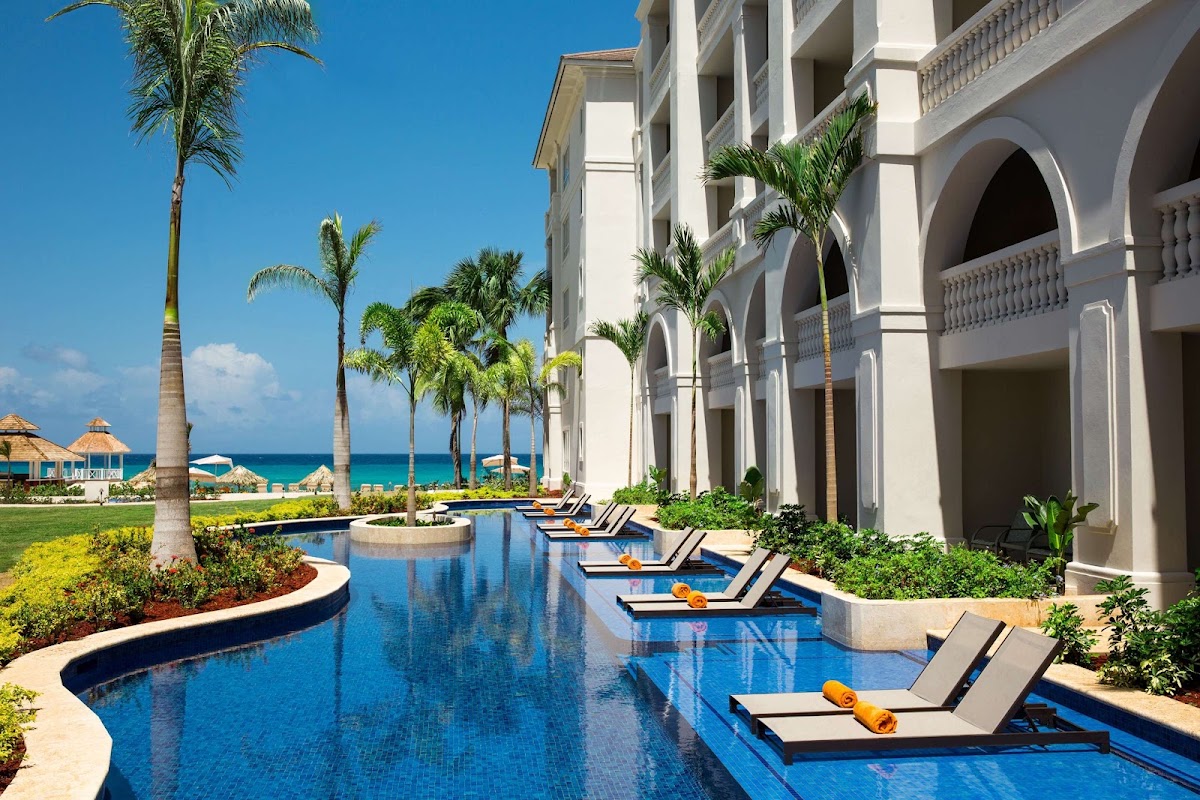 White resort building with arched columns overlooks a long blue pool lined with lounge chairs, palm trees, and the Caribbean Sea beyond.