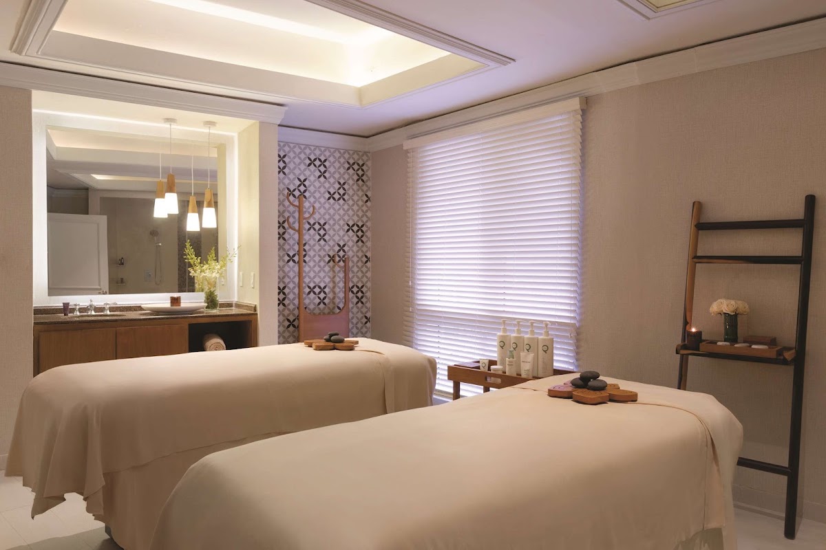 Two massage tables with cream draping sit in a spa treatment room with illuminated mirror, wooden vanity, and decorative shelving.
