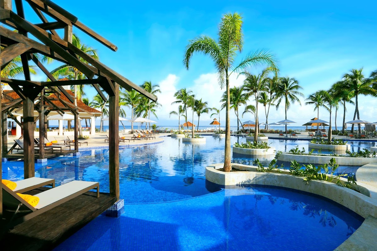 Hyatt Ziva Rose Hall pool area with wooden pergolas, loungers, palm trees, and ocean view beyond.