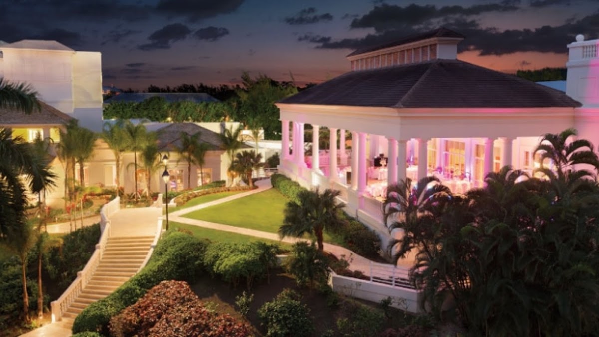 Hyatt Ziva Rose Hall illuminated at dusk with pink and white lighting, manicured lawn, palm trees, and colonial-style architecture against a twilight sky.