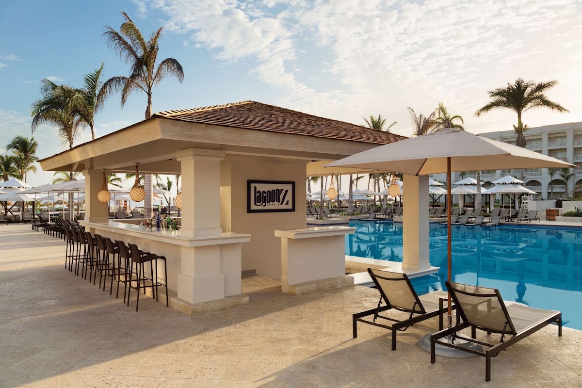 Beige poolside bar with black bar stools and Lagoon7 signage overlooks a bright blue swimming pool, with lounge chairs and palm trees nearby.