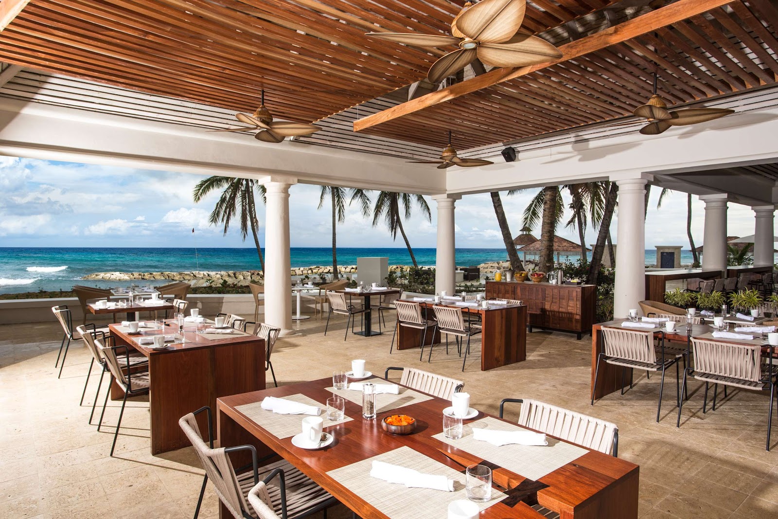 Beachfront dining pavilion at Hyatt Ziva Rose Hall with wooden tables, striped chairs, and ocean views between palm trees.