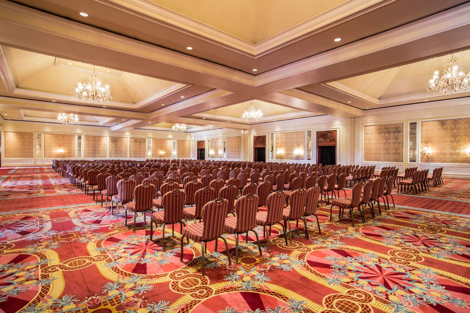 Large ballroom with burgundy striped chairs arranged in rows on ornate red and gold patterned carpet, coffered ceiling with crystal chandeliers and recessed lighting.