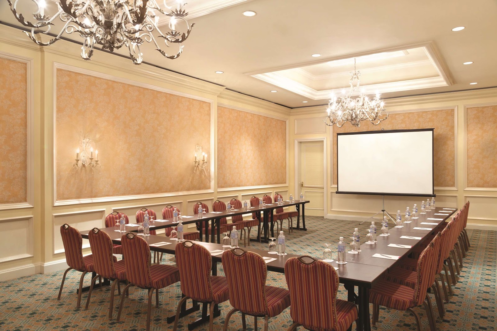 Elegant conference room with U-shaped table setup, red striped chairs, crystal chandeliers, and projection screen at Hyatt Ziva Rose Hall.