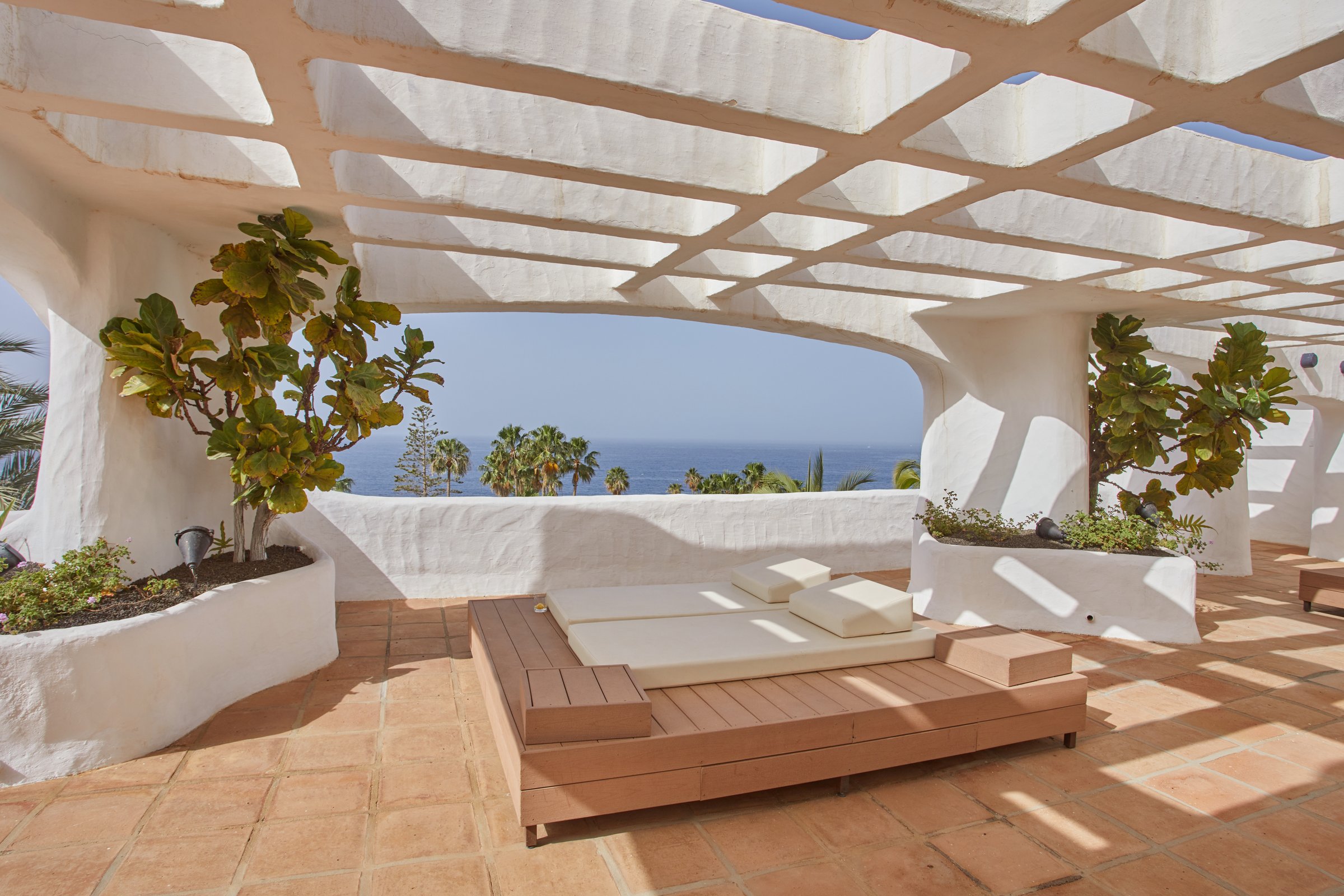 Sunny white terrace with a wooden daybed under a pergola, looking to palms and the ocean.