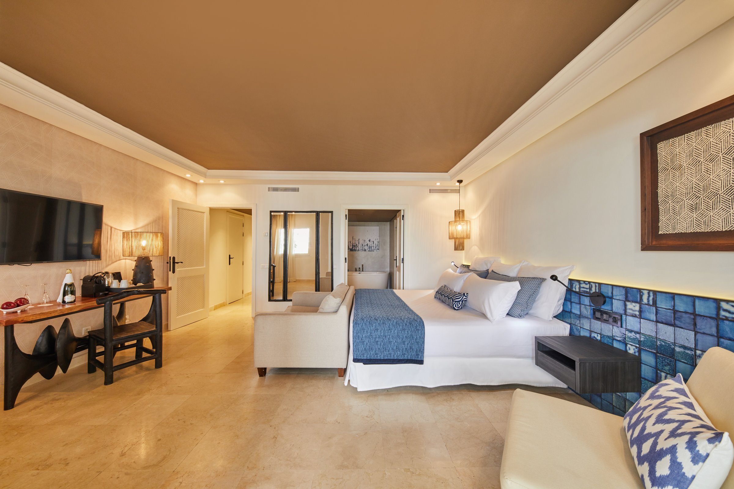Suite with blue-tiled bed wall, seating, welcome champagne and fruit, and a soaking tub beyond.
