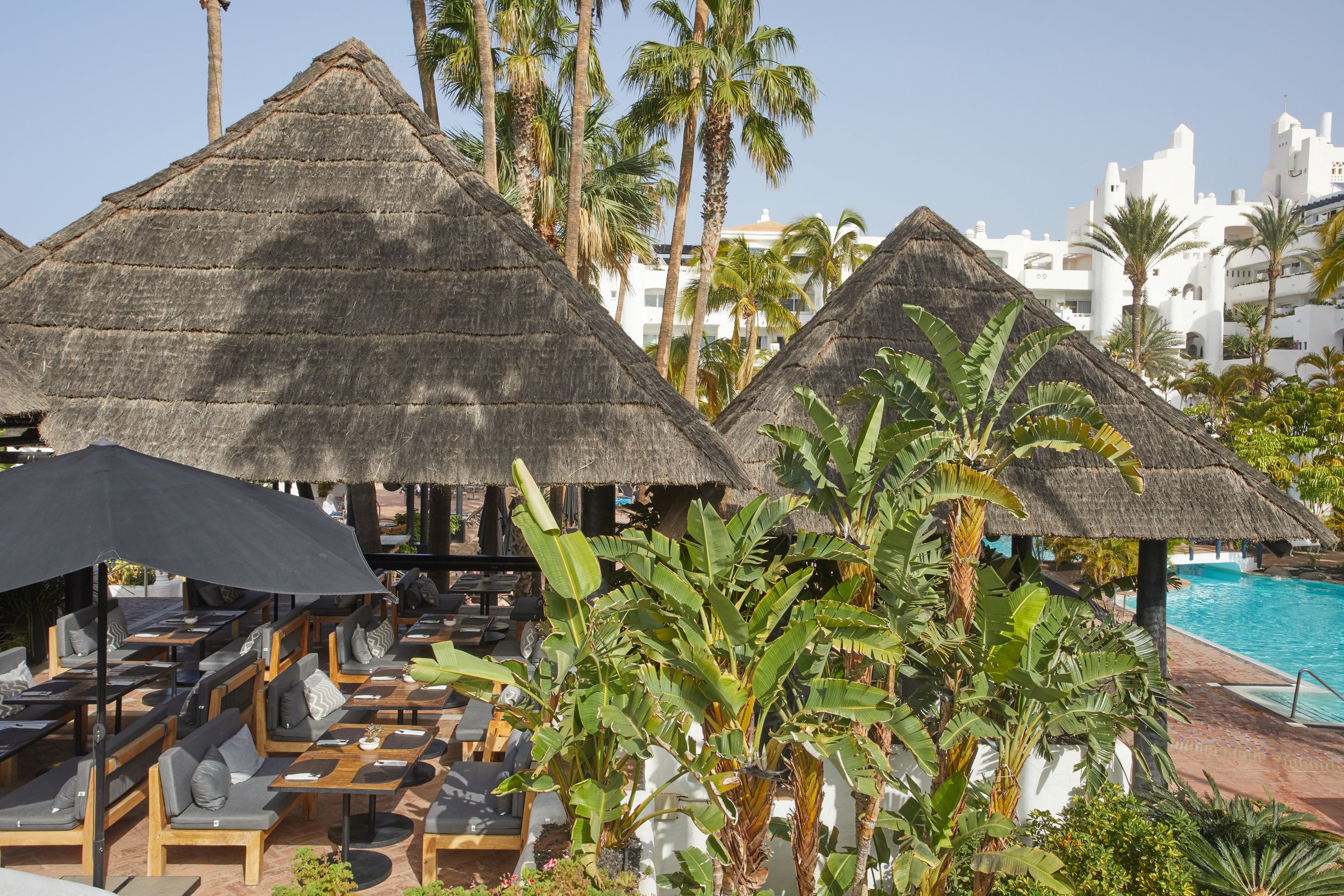 Barefoot Grill seating under thatched palapas with tropical plants and the white buildings behind.
