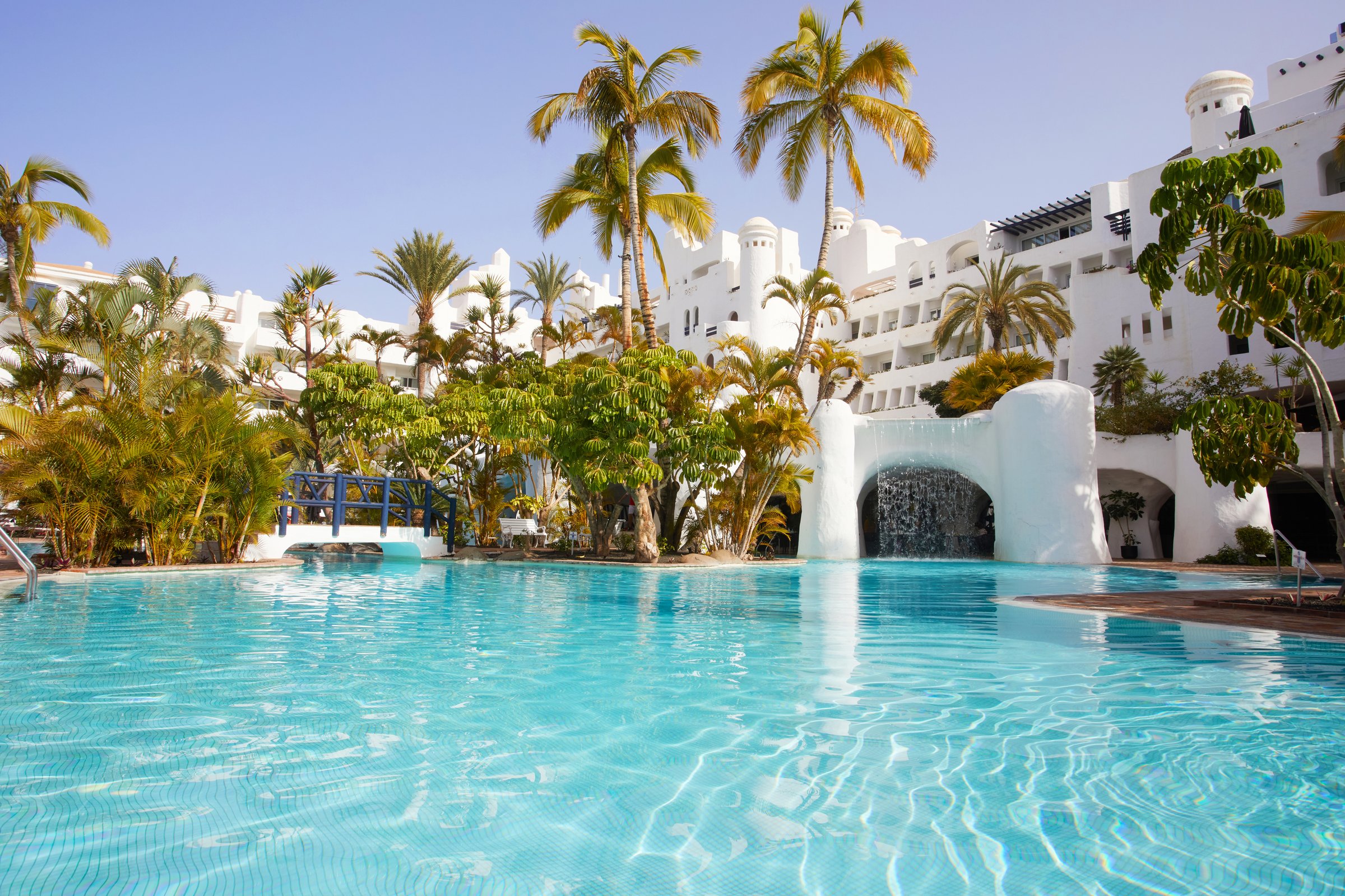 Bright turquoise pool with the little blue bridge, palms, waterfall and white turreted buildings.