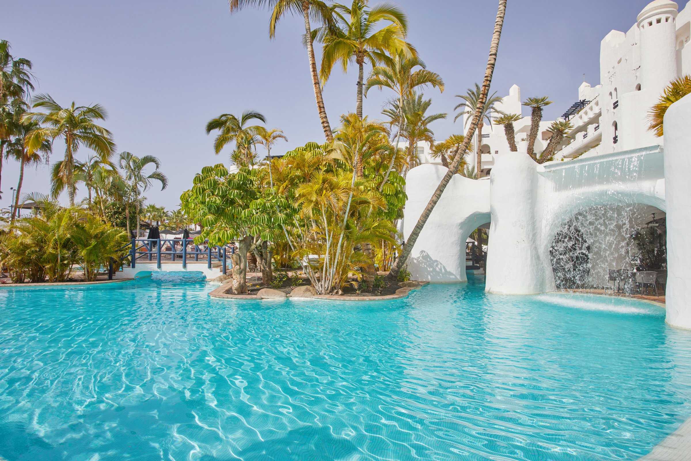 Sparkling pool wrapped around palm islands, with the white waterfall wall and shaded cafe tables.