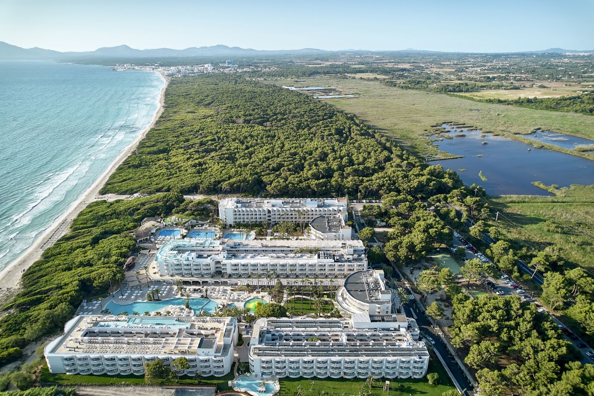 Aerial view of Iberostar Selection Albufera Playa with white buildings, pools, and beach surrounded by green wetlands and coastal town beyond.
