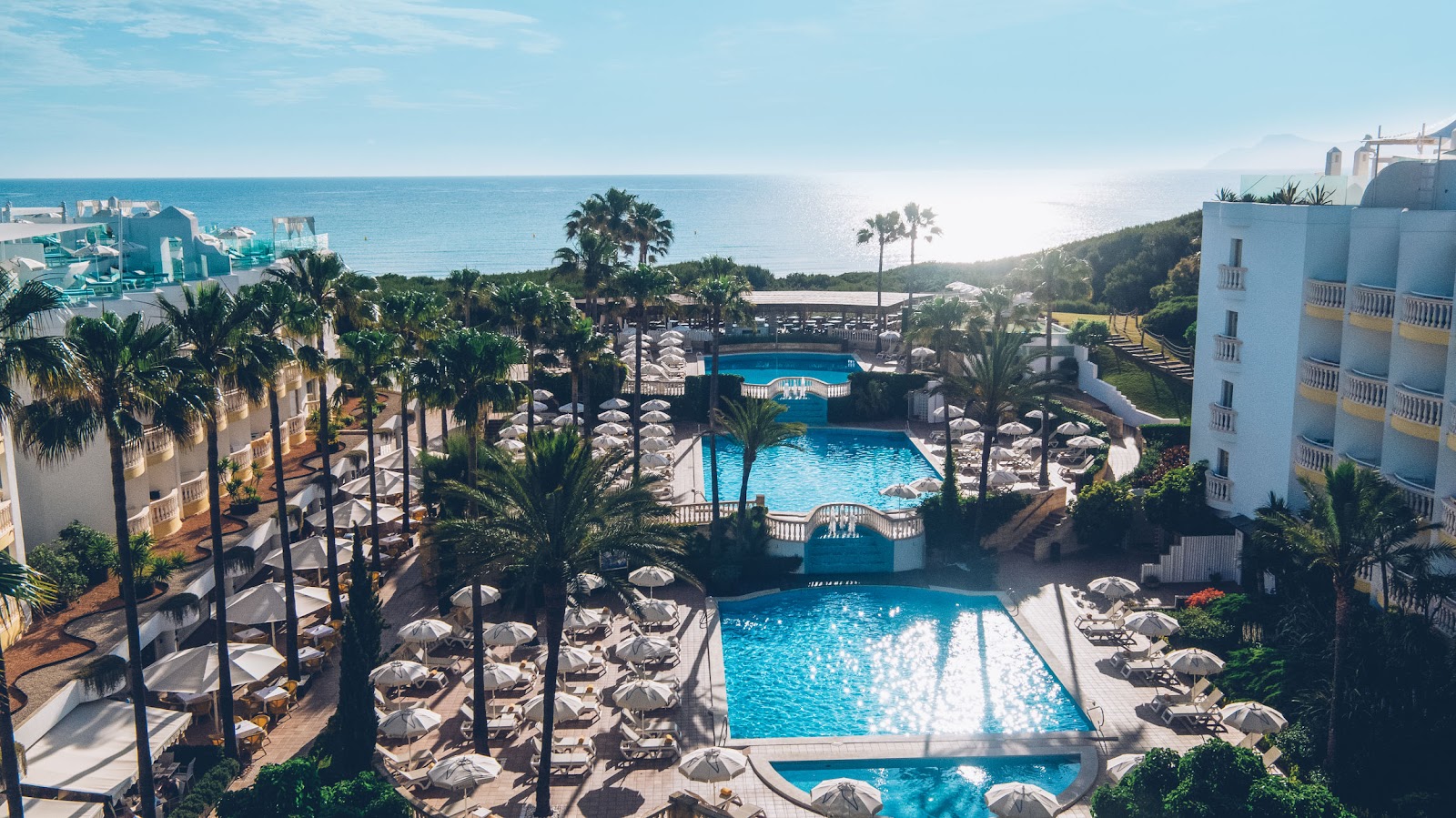 Aerial view of resort pools surrounded by palm trees, white umbrellas, and loungers, with Mediterranean Sea and coastal cliffs beyond.