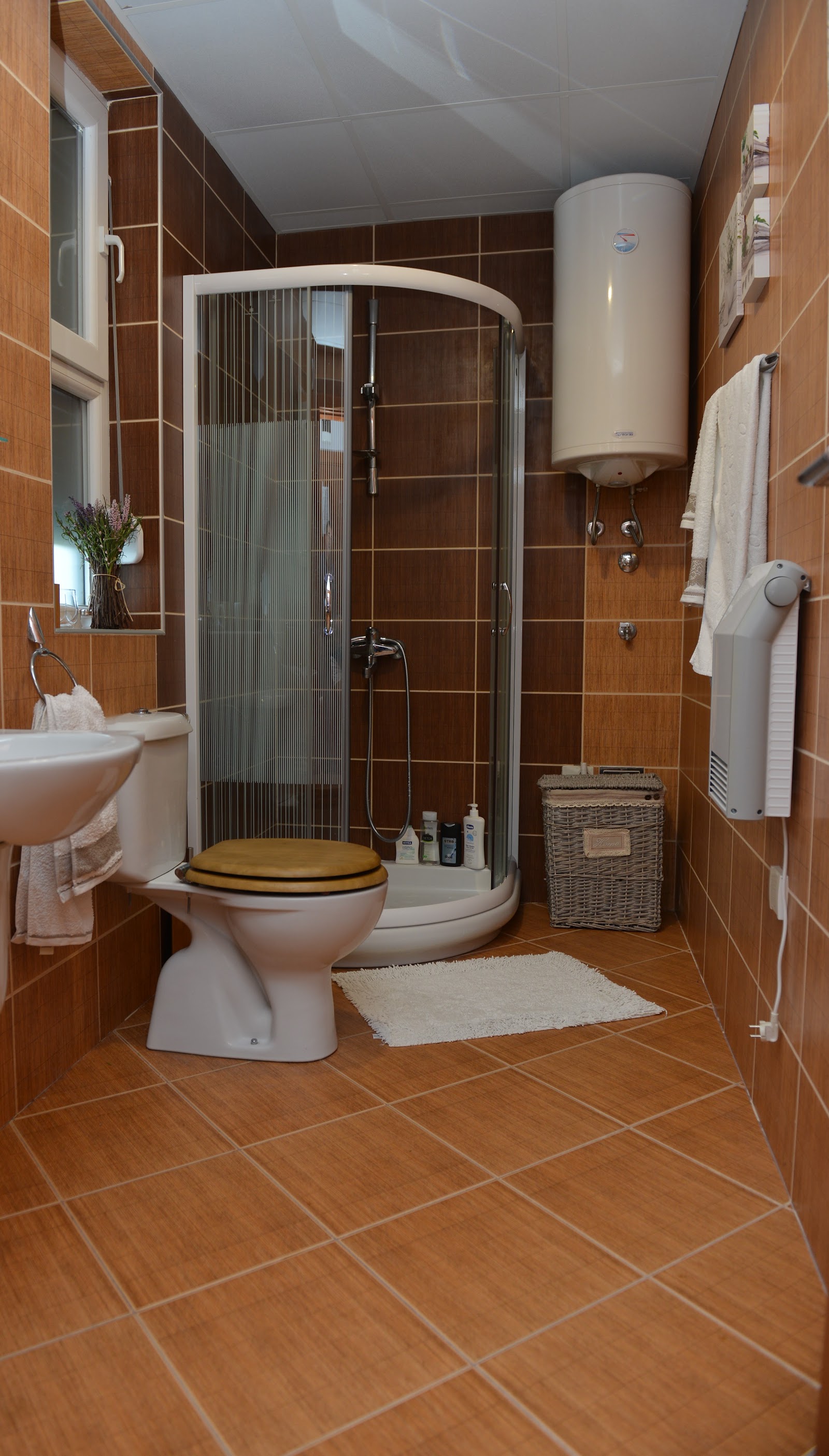Bathroom with brown tile walls, curved glass shower enclosure, toilet with yellow seat, white water heater, sink, towel rack, and wicker storage basket.