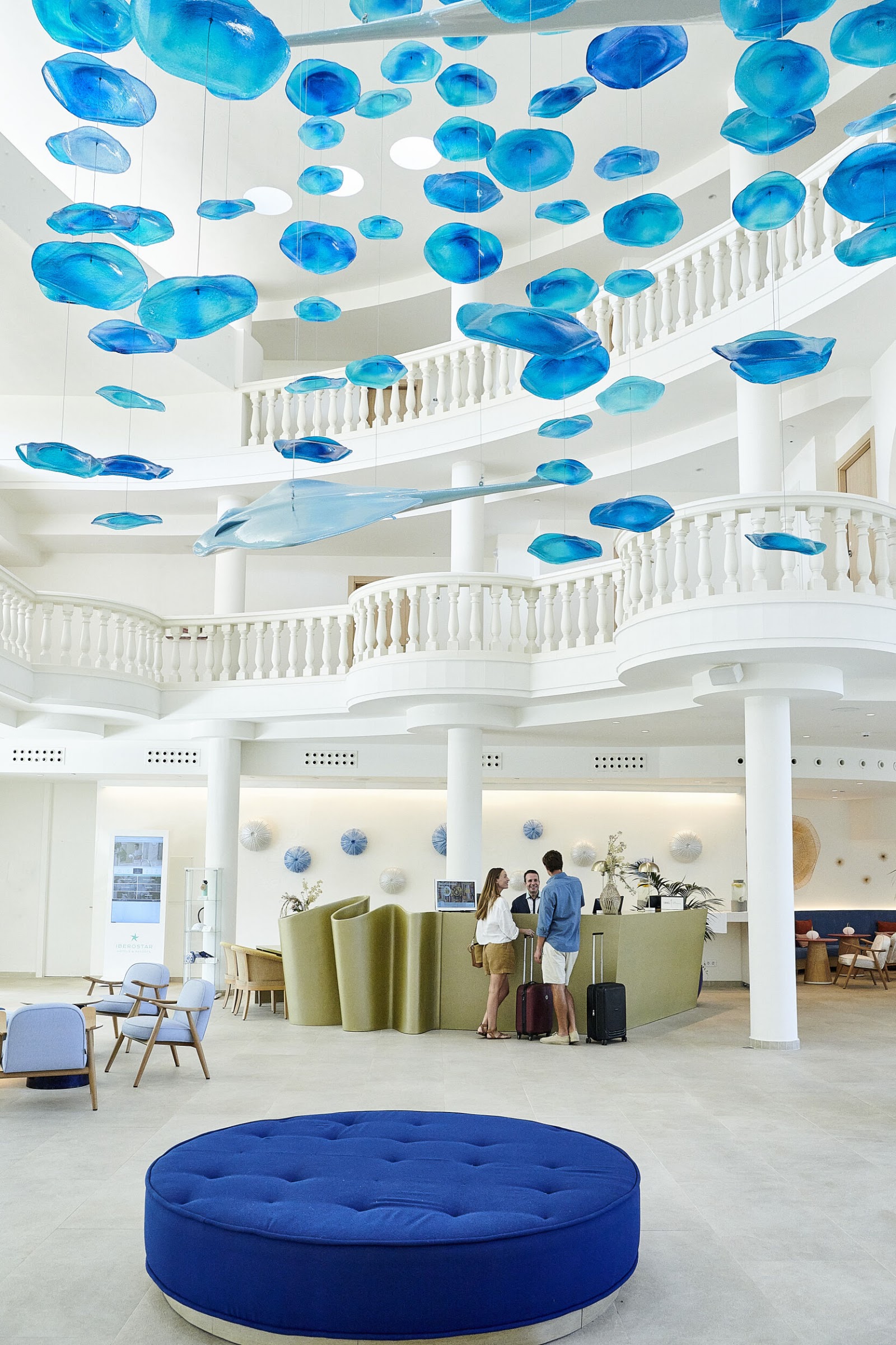 Modern lobby with blue glass fish sculptures hanging from white ceiling, curved balconies, and guests checking in at green reception desk.