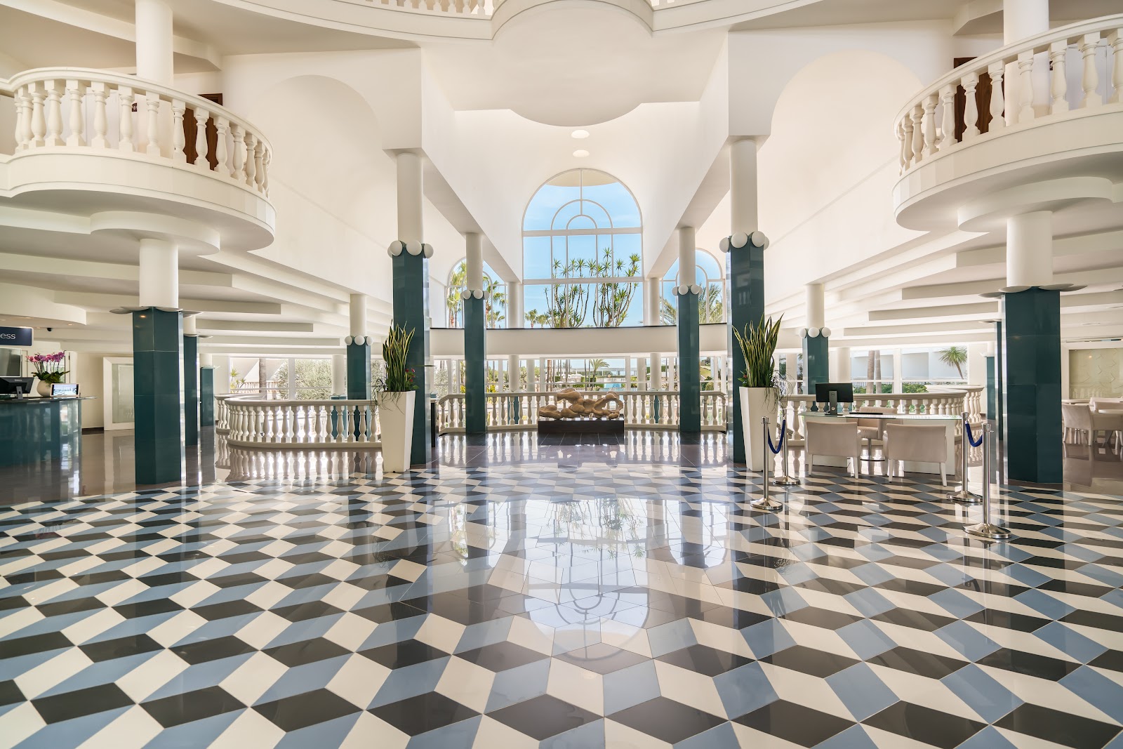 Grand lobby with diamond-patterned marble floor, curved balconies, green columns, and arched windows overlooking palm trees at Iberostar Selection Albufera Playa.