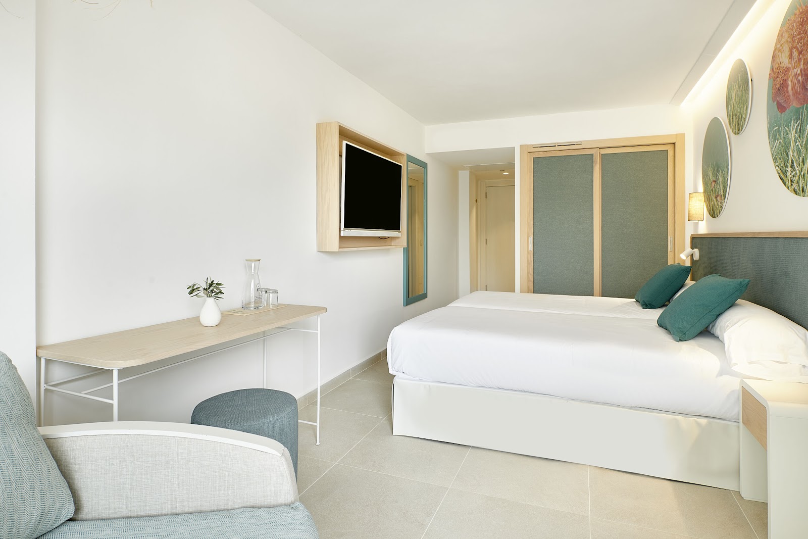 Modern guest room with white bed, teal accent pillows, desk with mounted TV, and light wood furnishings at Iberostar Selection Albufera Playa.