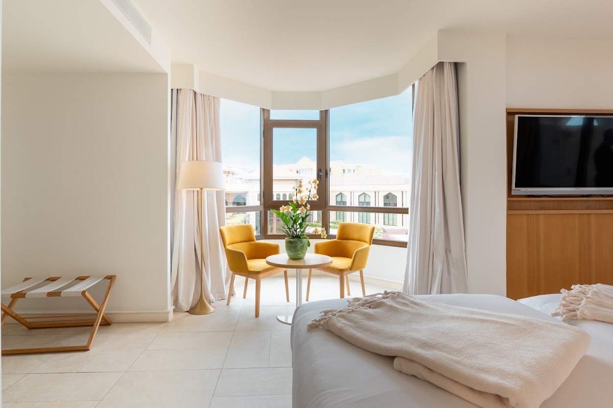 Spacious resort suite with bed, yellow armchairs, and balcony overlooking coastal town buildings under blue sky.