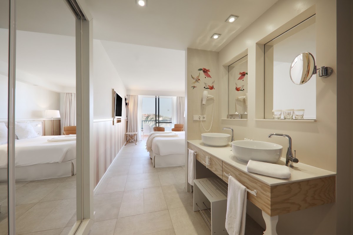 Modern bathroom with dual vessel sinks, round mirrors, and floral wall art, adjoining a bedroom with ocean-view balcony at Iberostar Selection Anthelia.
