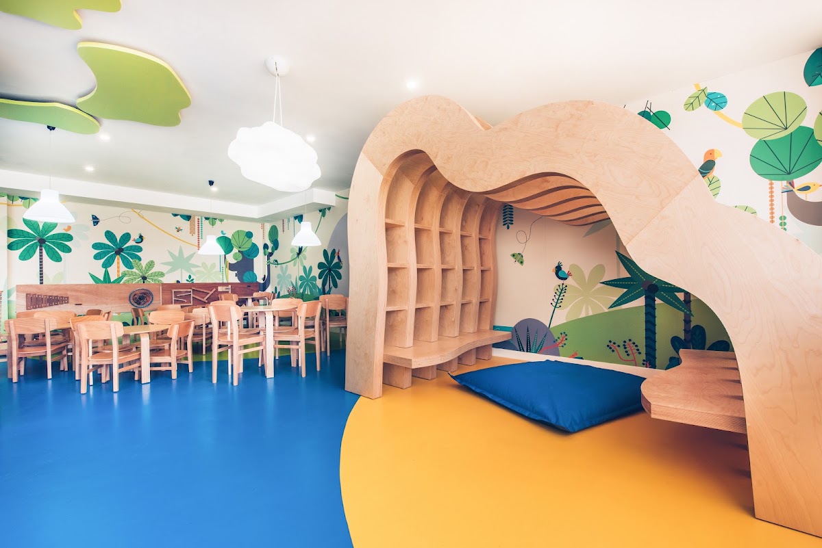 Kids club interior with wooden wave-shaped structure, blue cushion, tropical wall murals, and dining tables in the background.