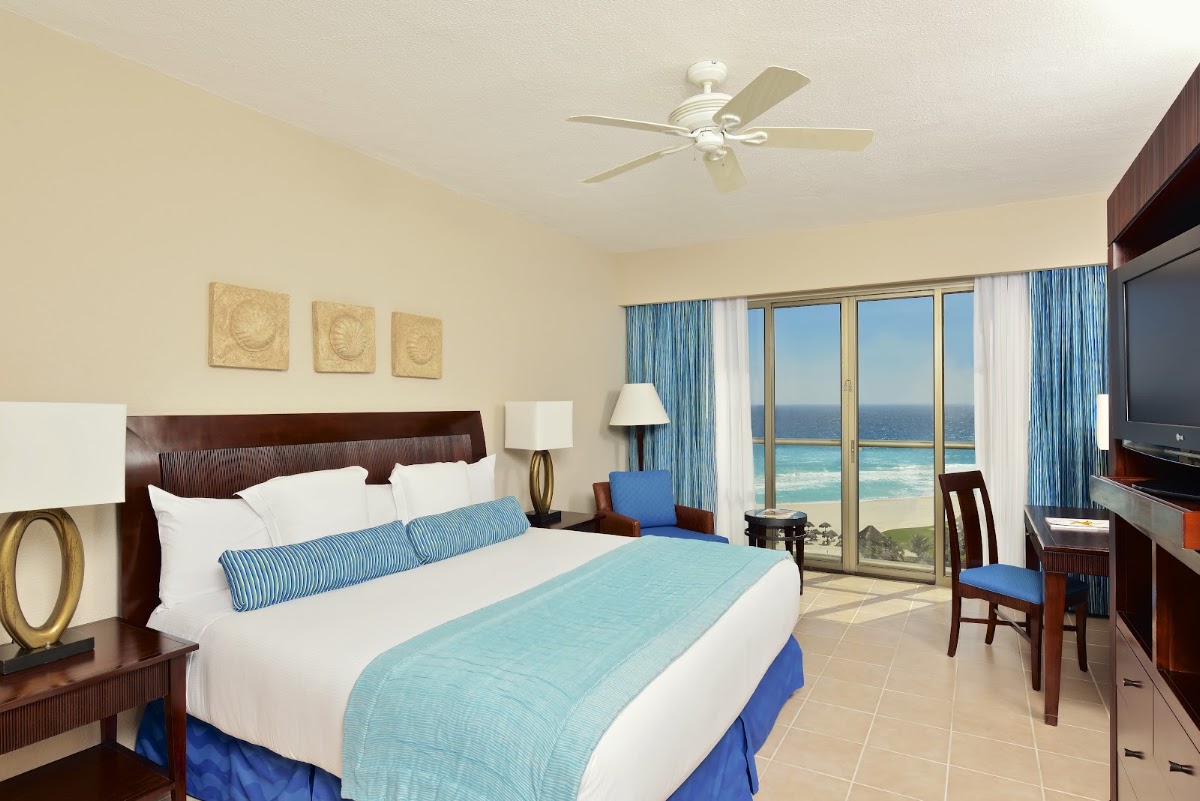 Beachfront resort bedroom with ocean view through floor-to-ceiling windows, queen bed with white linens, dark wood furniture, and ceiling fan.