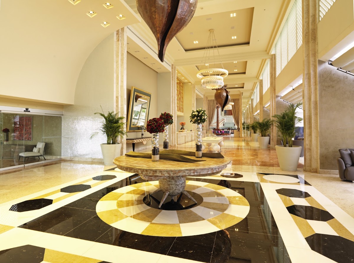 Elegant lobby with geometric black and gold floor pattern, central round table with wooden sculpture overhead, crystal chandelier, and potted plants lining the hallway.