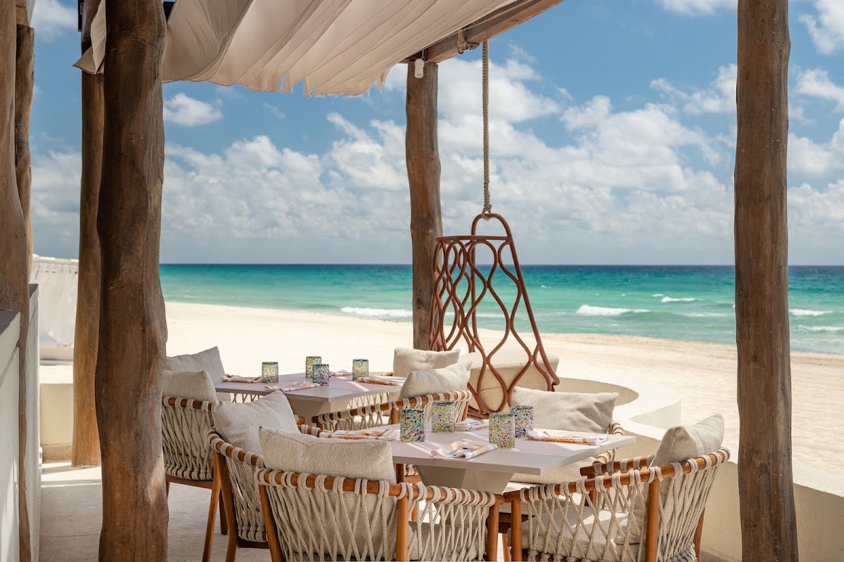 Beachfront dining pavilion with woven chairs, tables set with glasses and place settings, a hanging rope swing, wooden posts, canvas shade, and turquoise ocean beyond white sand.