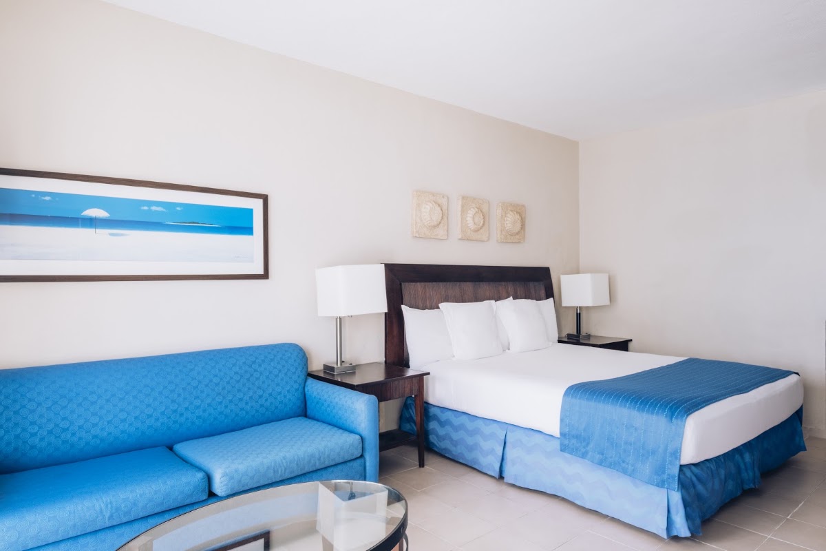 Resort guest room with blue sofa, queen bed with white bedding, dark wood headboard, and landscape artwork on beige walls.