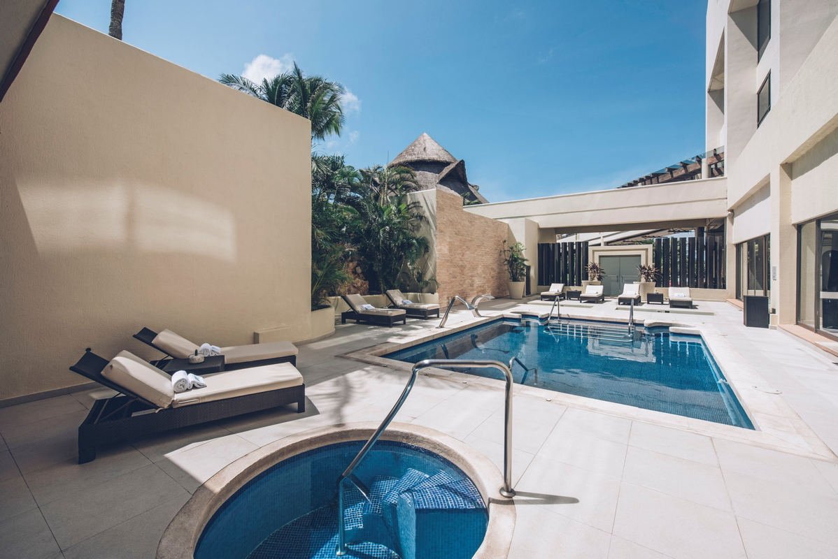 Resort pool deck with lounge chairs, hot tub in foreground, main swimming pool, thatched-roof structure, and white building with covered seating area under clear sky.