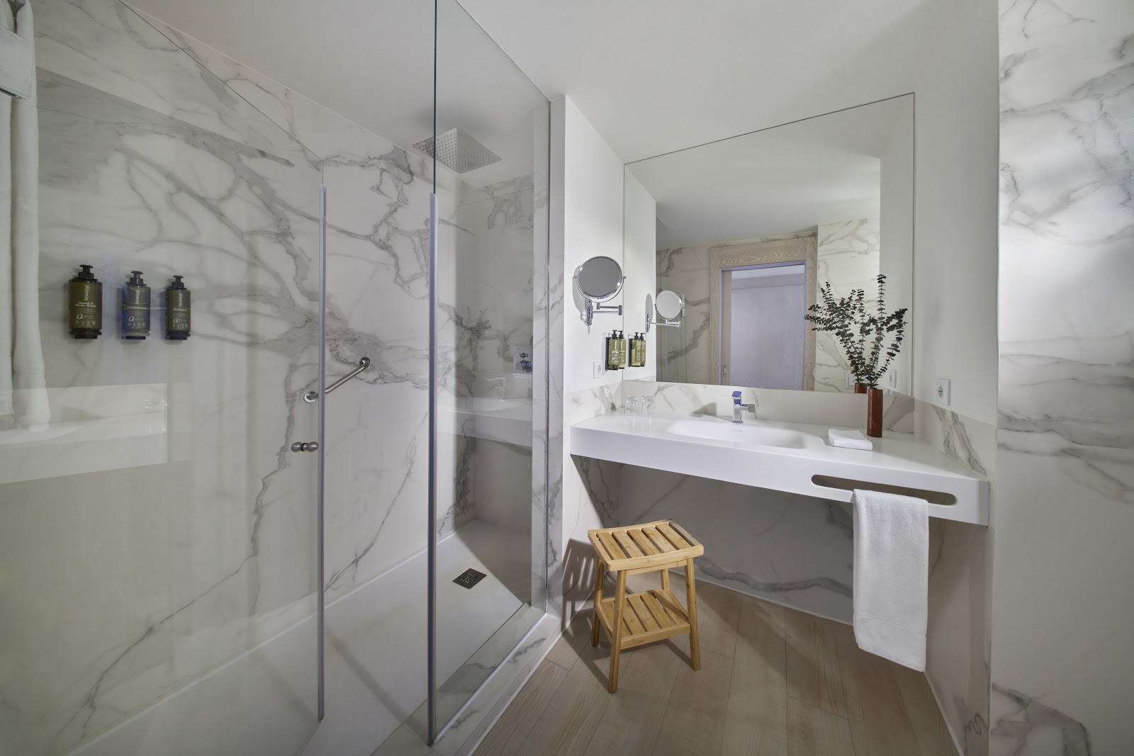 Modern bathroom with marble walls, glass shower enclosure, white vanity with large mirror, and wooden stool at Iberostar Selection Fuerteventura Palace.