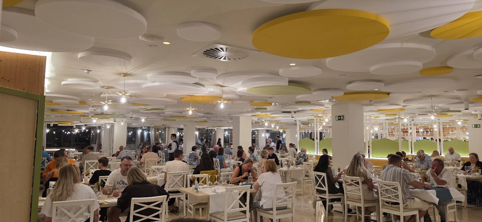 Dining room at Iberostar Selection Fuerteventura Palace with white cross-back chairs, round tables, and yellow-accented coffered ceiling, guests dining in evening light.