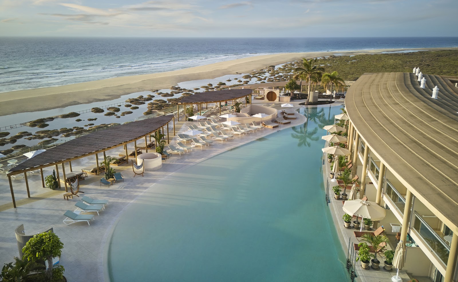 Iberostar Selection Fuerteventura Palace overlooks a curved lagoon pool with loungers, beach, and ocean beyond dotted with palm trees.