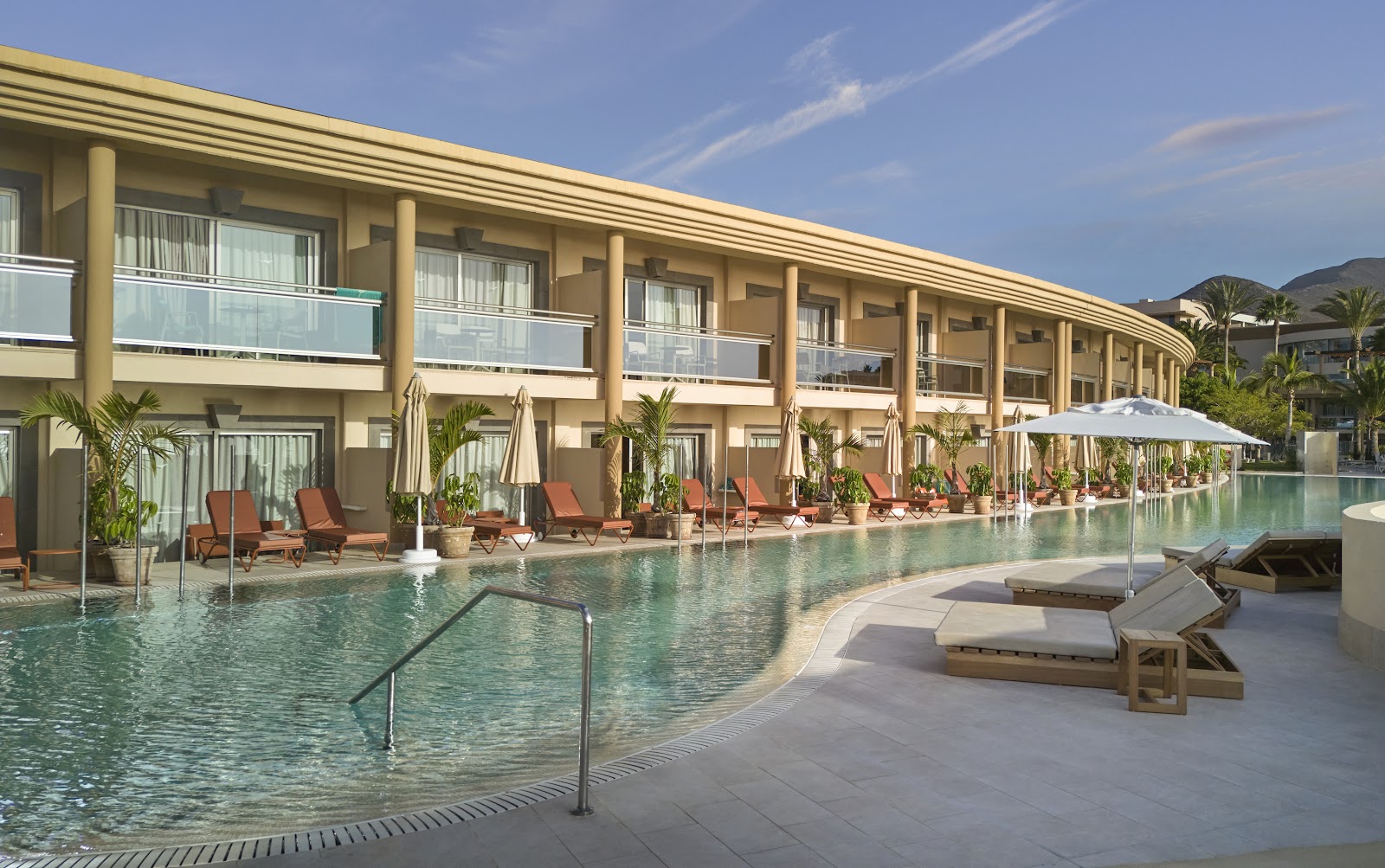 Iberostar Selection Fuerteventura Palace pool area with tan building, loungers, umbrellas, and palm trees under clear sky.