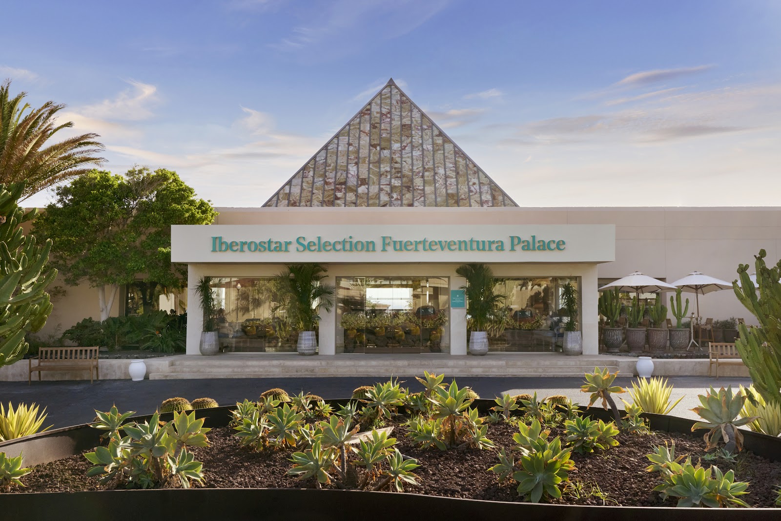 Iberostar Selection Fuerteventura Palace entrance with pyramidal glass roof, white facade, desert plants, and palm trees under blue sky.