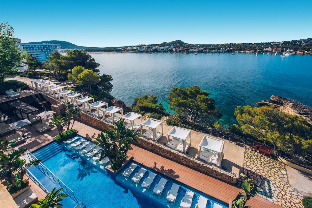 Resort pool and white cabanas overlook a turquoise bay with sailboats, pine trees, and hillside homes across the water.